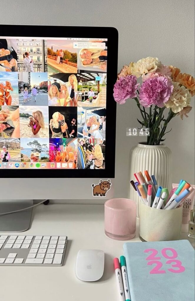 Workspace with computer displaying photos, 2023 planner, colorful pens, and flowers in vase, creating a vibrant, organized desk setup.