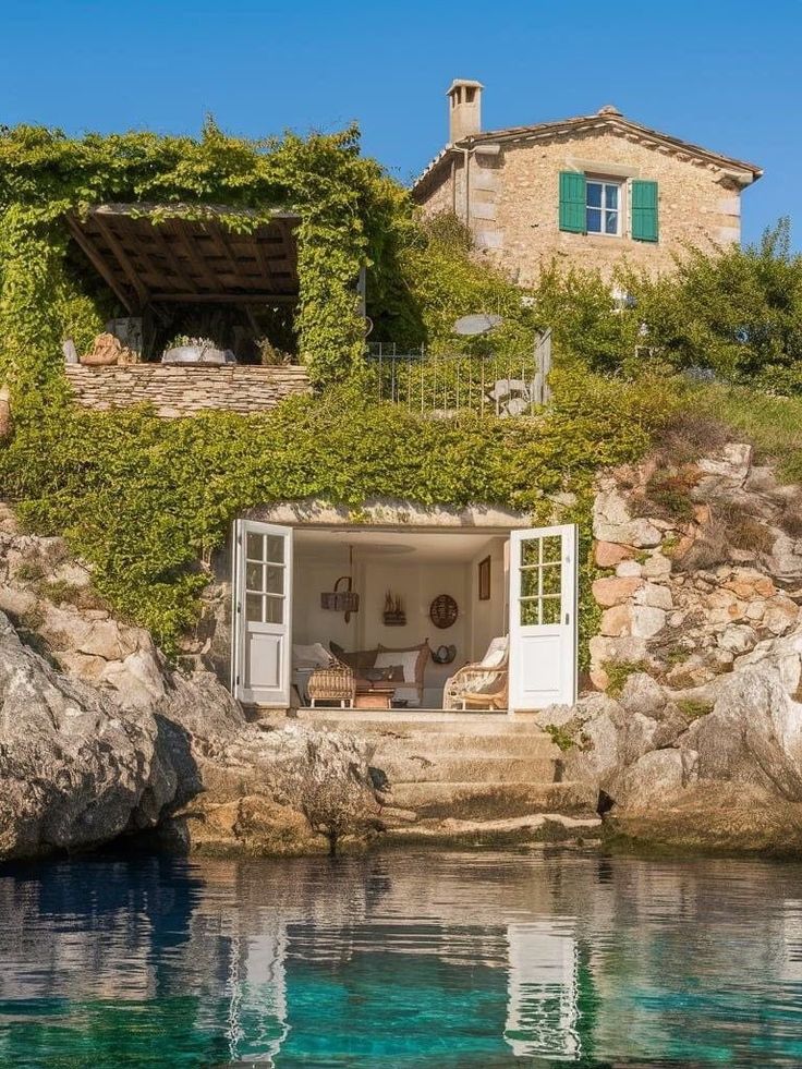 Stone house with lush greenery overlooks turquoise waters, featuring a cozy open room with white doors.
