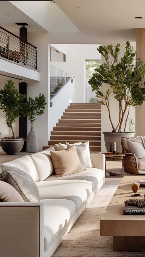 Modern living room with stylish beige sofa, wooden coffee table, and indoor plants near a staircase with glass railing.