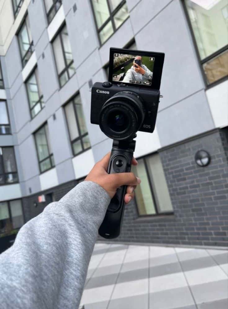 3a7c5610-f0b2-4afd-b9ab-5873e6a052bf - Handy Home Person holding Canon camera with flip screen, capturing a selfie in front of modern building.