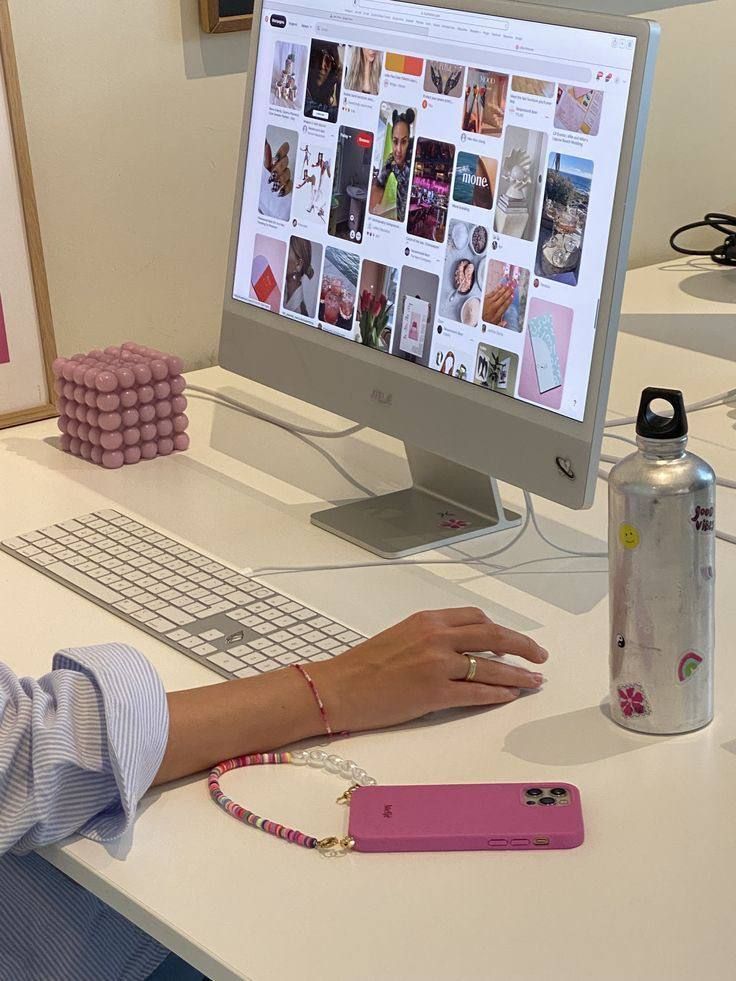Person browsing Pinterest on a desktop with a colorful phone and water bottle nearby on a white desk in an office setting.
