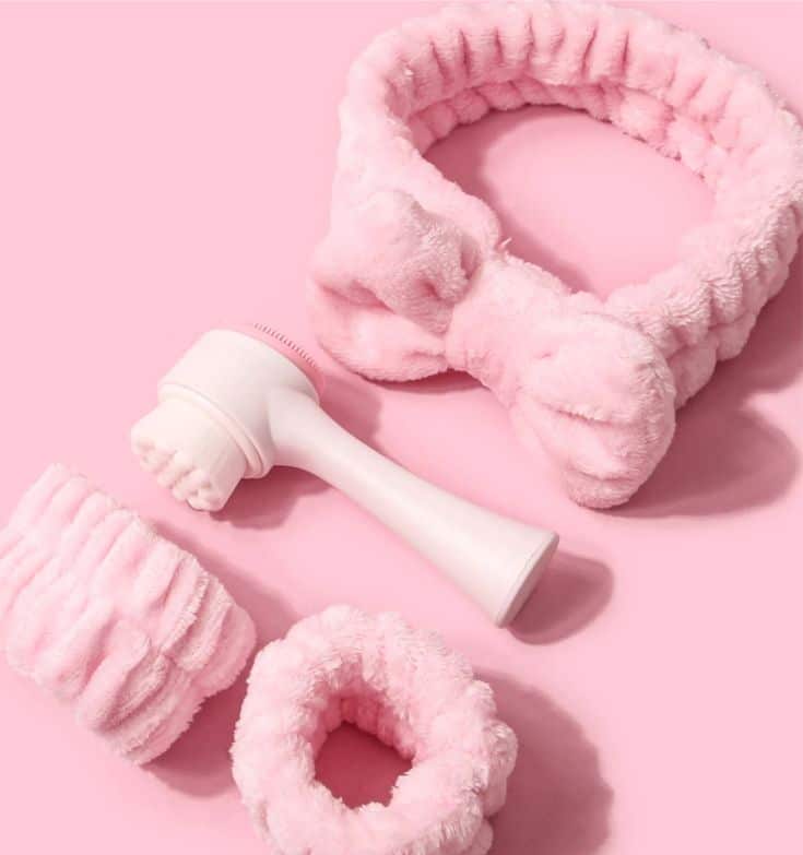 Soft pink spa accessories: headband, wristbands, and facial cleansing brush on a matching pink background. Relaxation essentials.
