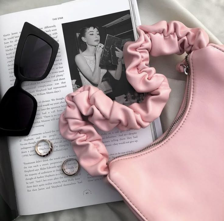 Pink scrunchie bag and sunglasses on open book, with rings, creating a chic and stylish flatlay.
