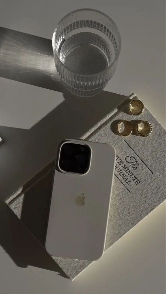 Minimalist scene with a smartphone, glass of water, a journal, and gold rings on a table, bathed in soft light.