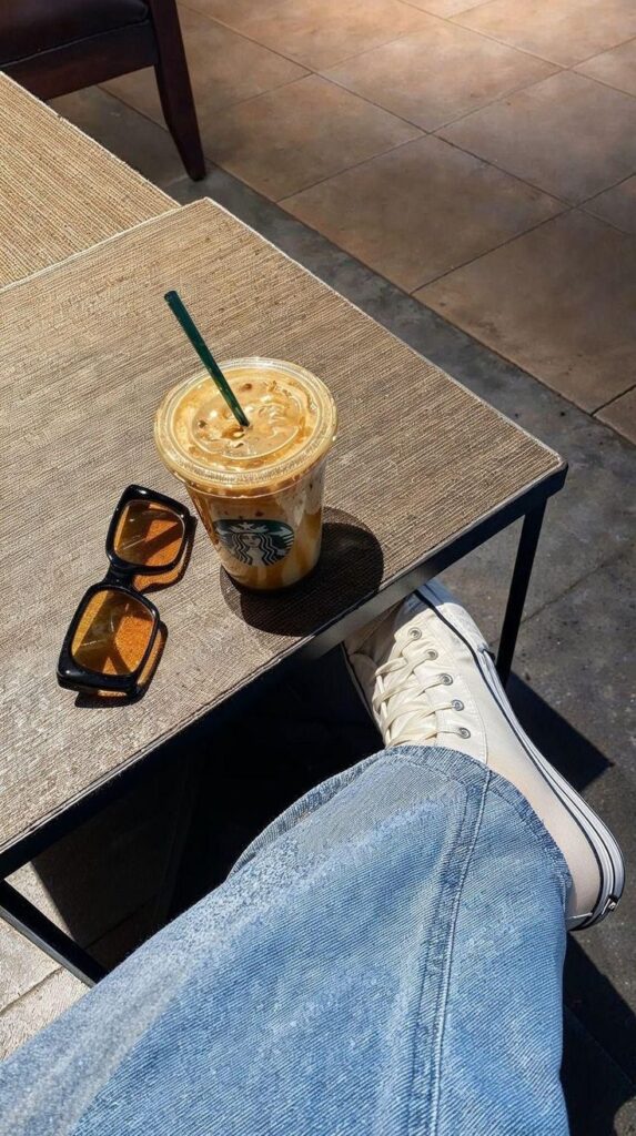 Casual cafe moment: iced coffee with green straw, sunglasses, and white sneakers on a sunny day. Relaxed vibe.