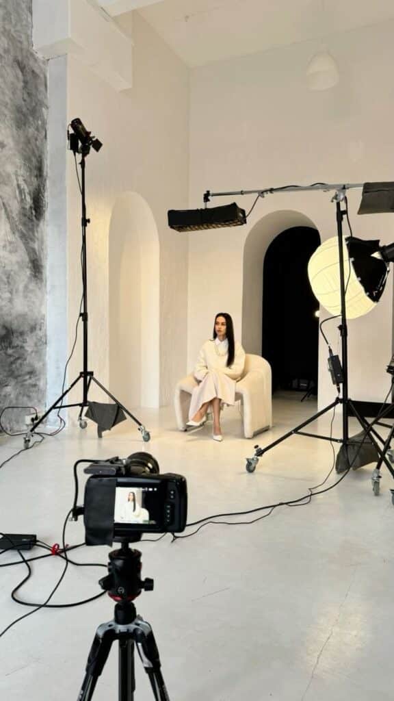 Photography studio setup with professional lighting, camera, and model sitting on a chair in a minimalist setting.
