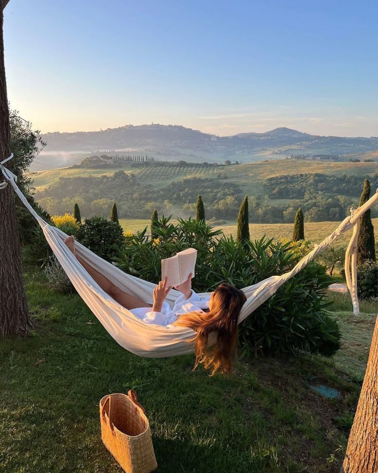 Woman relaxing in hammock reading a book with scenic countryside view; peaceful outdoor retreat.