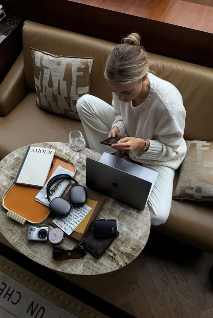 Woman in a cozy setting, working on a laptop and phone, surrounded by books, headphones, and accessories on a marble table.