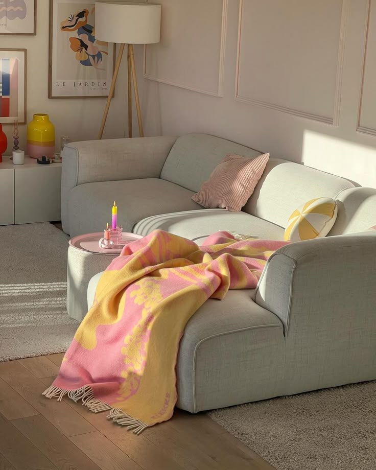 Cozy living room with a light gray sofa, colorful throw, pink pillow, and art on the walls, basking in warm sunlight.
