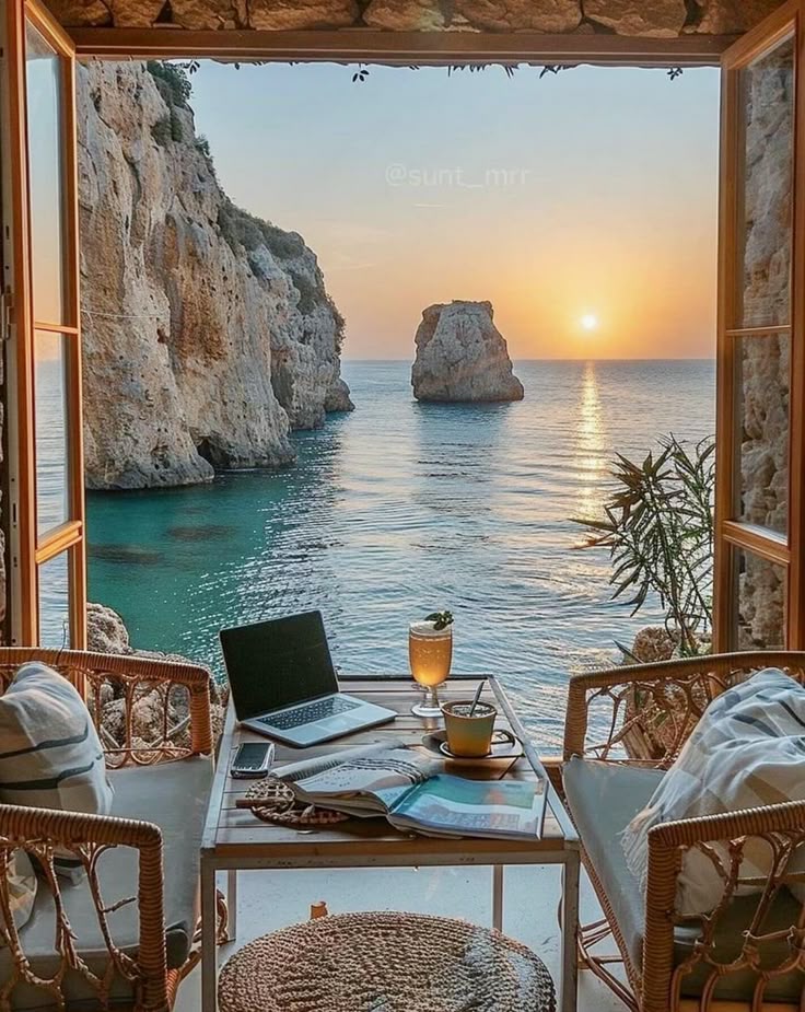Coastal retreat, ocean view through open doors with sunset, cozy chairs, table, laptop, book, and drinks. Perfect work and relax spot.