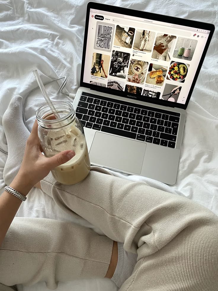Cozy setting with person holding iced coffee by a laptop displaying Pinterest on a bed. Relaxing at home lifestyle.