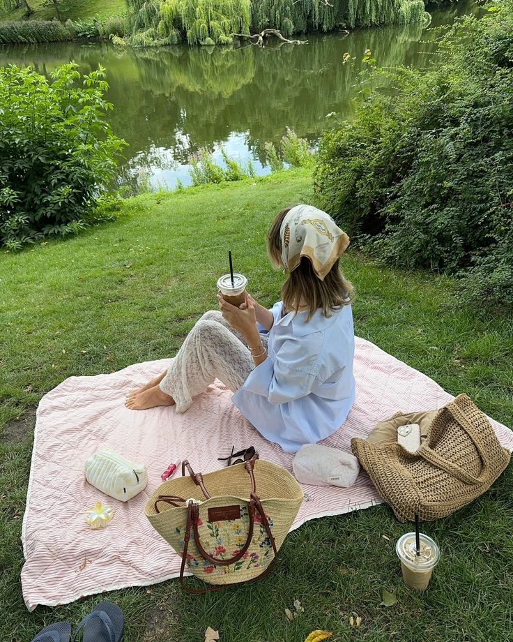 Woman relaxing by a lake on a picnic blanket, enjoying a cold drink amidst lush greenery.
