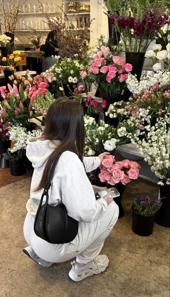 Woman in a floral shop surrounded by colorful bouquets, holding a smartphone, wearing a white hoodie and sneakers.