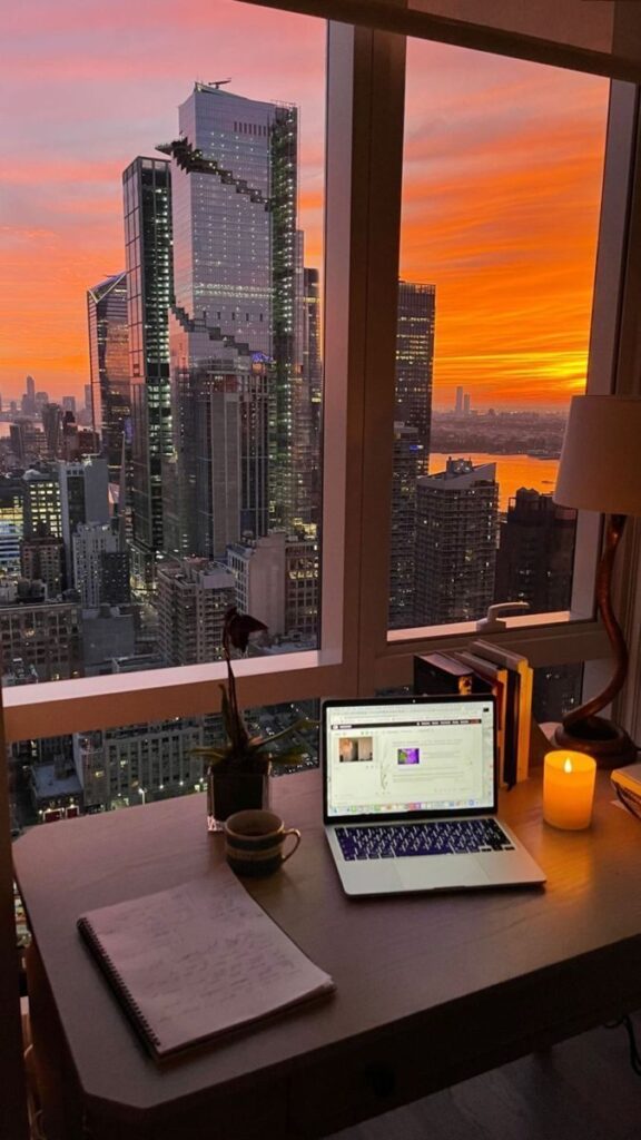 Cozy home office with a laptop, candle, and cityscape view at sunset, creating a productive and inspirational work environment.