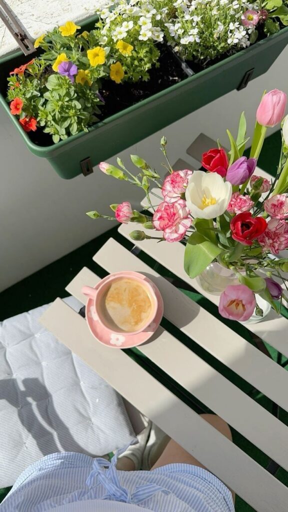 Cozy balcony scene with fresh flowers, a pink coffee cup, and vibrant plants, perfect for a relaxing morning.
