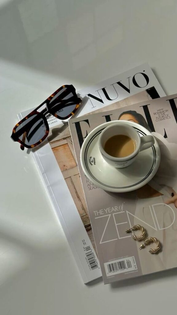 Cup of coffee on fashion magazines with sunglasses and earrings nearby on a sunlit table.
