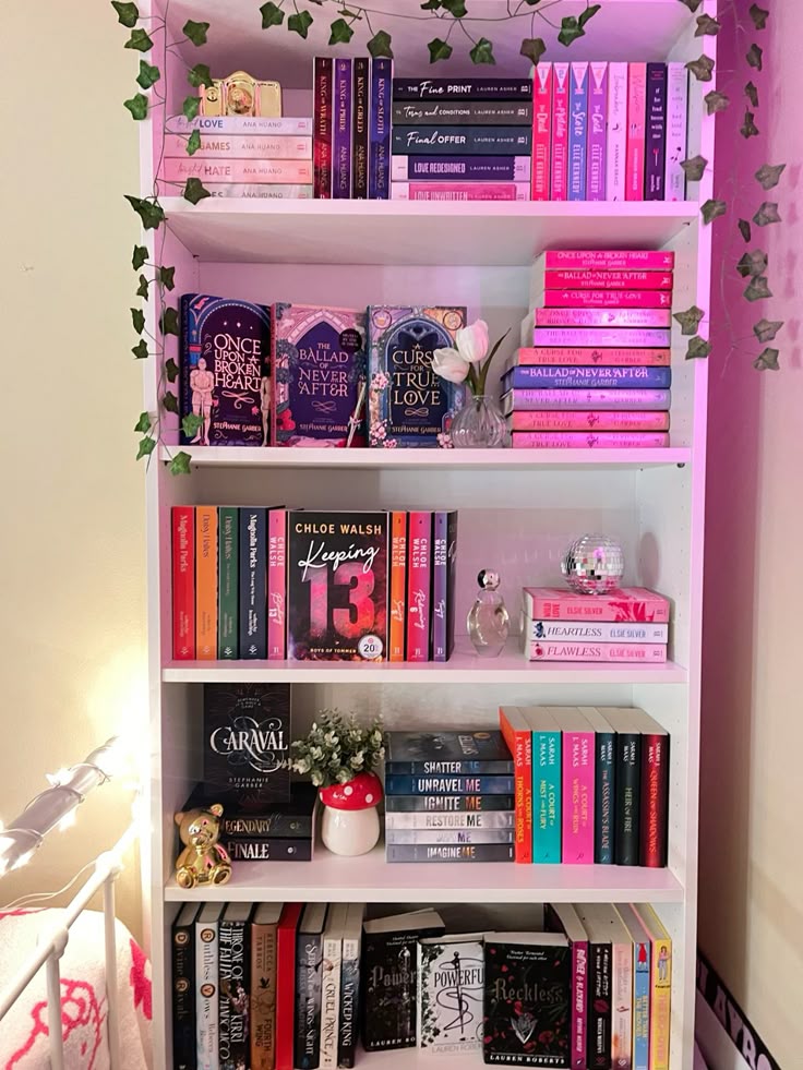 Colorful bookshelf with novels, decorative plants, and figurines. Aesthetic book arrangement with pink and white tones.