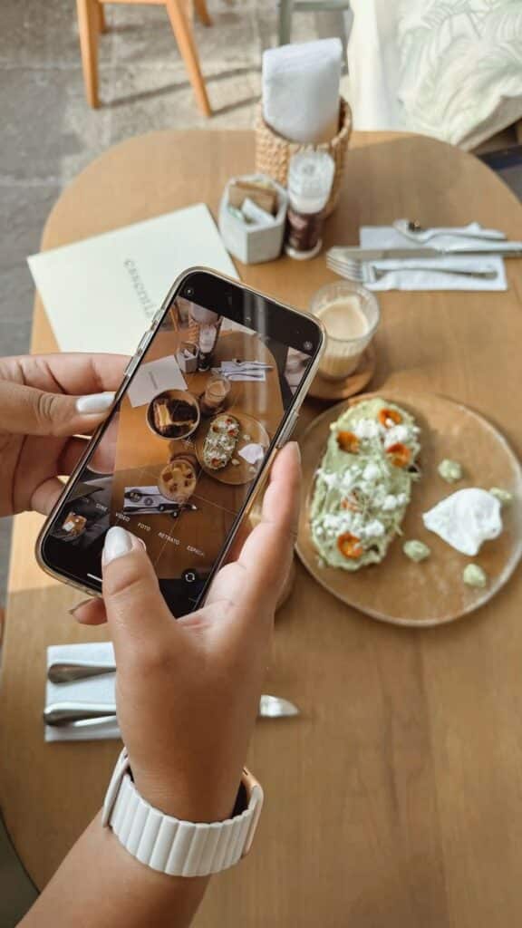 Person capturing a brunch table setting with phone, featuring dishes and a latte on a wooden table for Instagram.