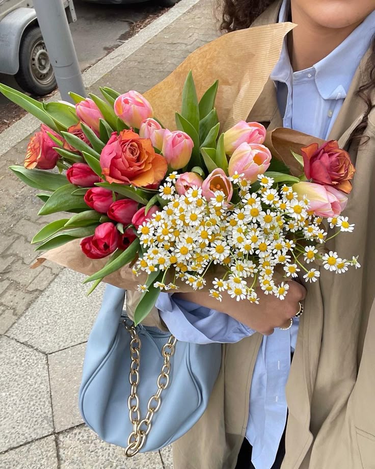 Person holding a vibrant bouquet of roses, tulips, daisies, paired with a light blue purse on a city sidewalk.