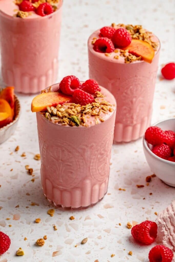 Delicious pink smoothies topped with fresh raspberries, peach slices, and granola, served in elegant glasses on a marble surface.