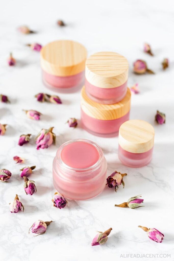 Pink homemade lip balms in glass jars with wooden lids surrounded by dried rosebuds on a marble surface.