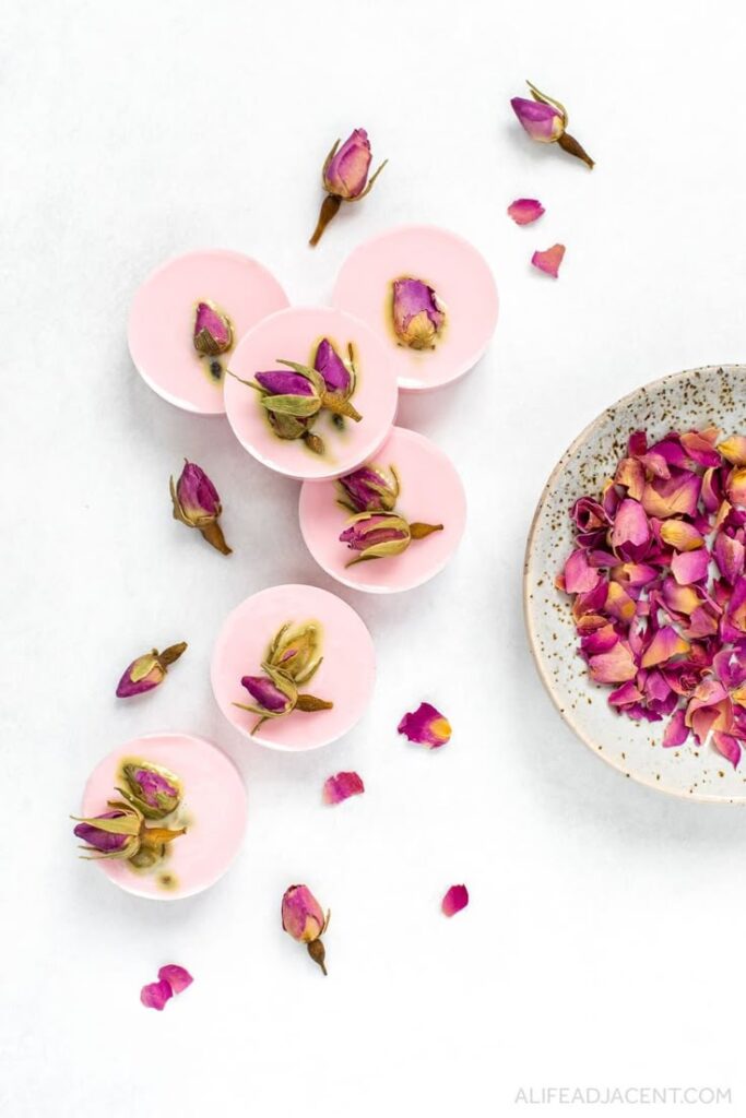 Pink rose petal wax melts with dried rosebuds on a white background, accompanied by a speckled bowl of petals. Elegant and soothing.