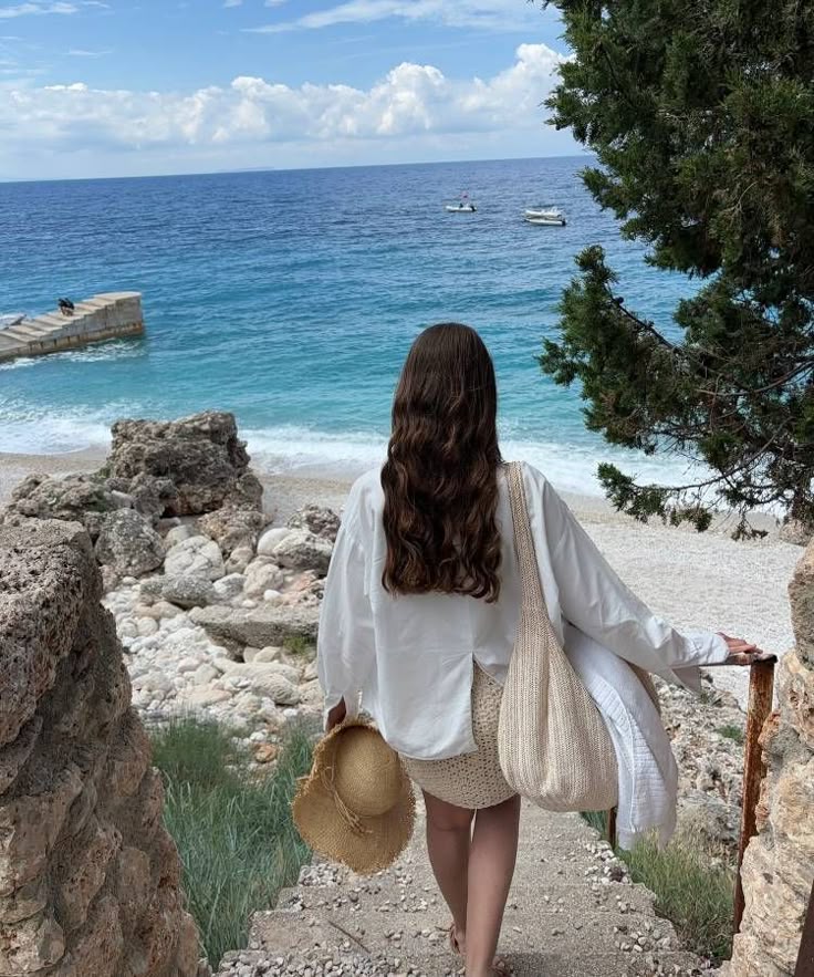 Woman in white walks down stone steps toward a sandy beach and blue ocean, carrying a hat and bag, surrounded by nature.
