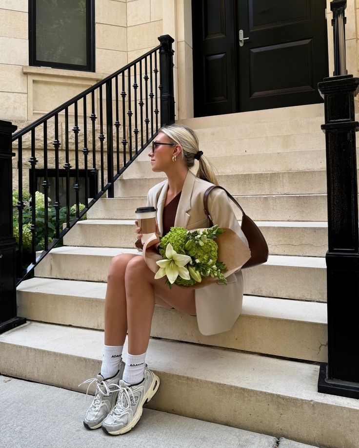 Woman in stylish blazer sits on steps with coffee and flowers, wearing sunglasses and sneakers. Urban chic fashion scene.