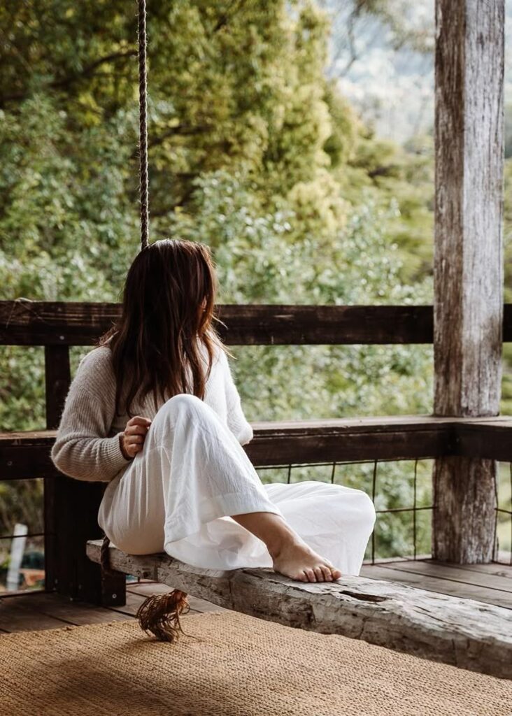 Woman in white dress sitting on wooden swing, enjoying serene forest view from porch. Peaceful nature retreat setting.