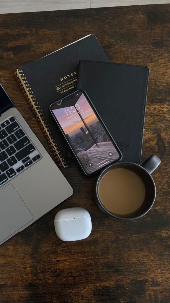 Laptop, smartphone, notebooks, earbuds, and coffee on a wooden desk, creating a productive workspace setup.