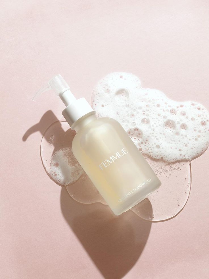 Transparent bottle of Femmue cleansing gel on a pink background with bubbly foam, casting a soft shadow, promoting skincare.