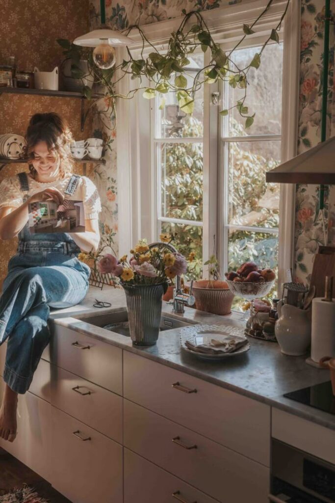 Cozy kitchen scene with a woman in overalls sitting by the window, surrounded by plants and flowers, reading a book.