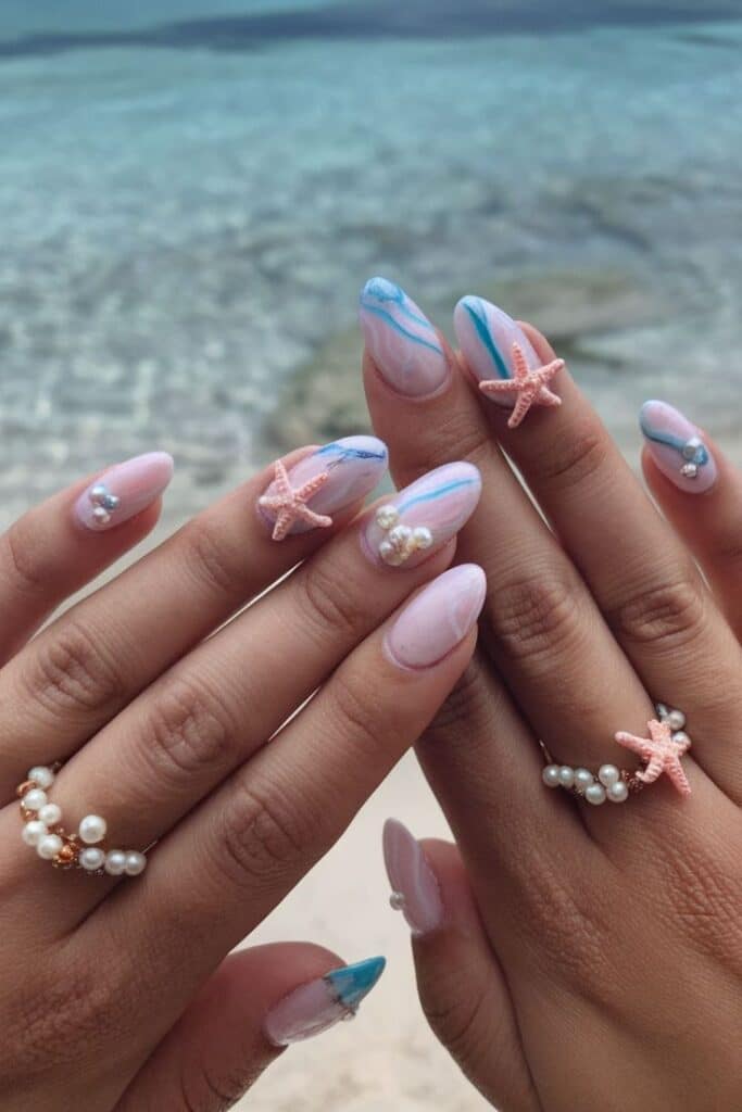 Beach-themed nail art with blue waves, starfish, and pearl accents, set against a serene ocean backdrop. Perfect summer manicure.