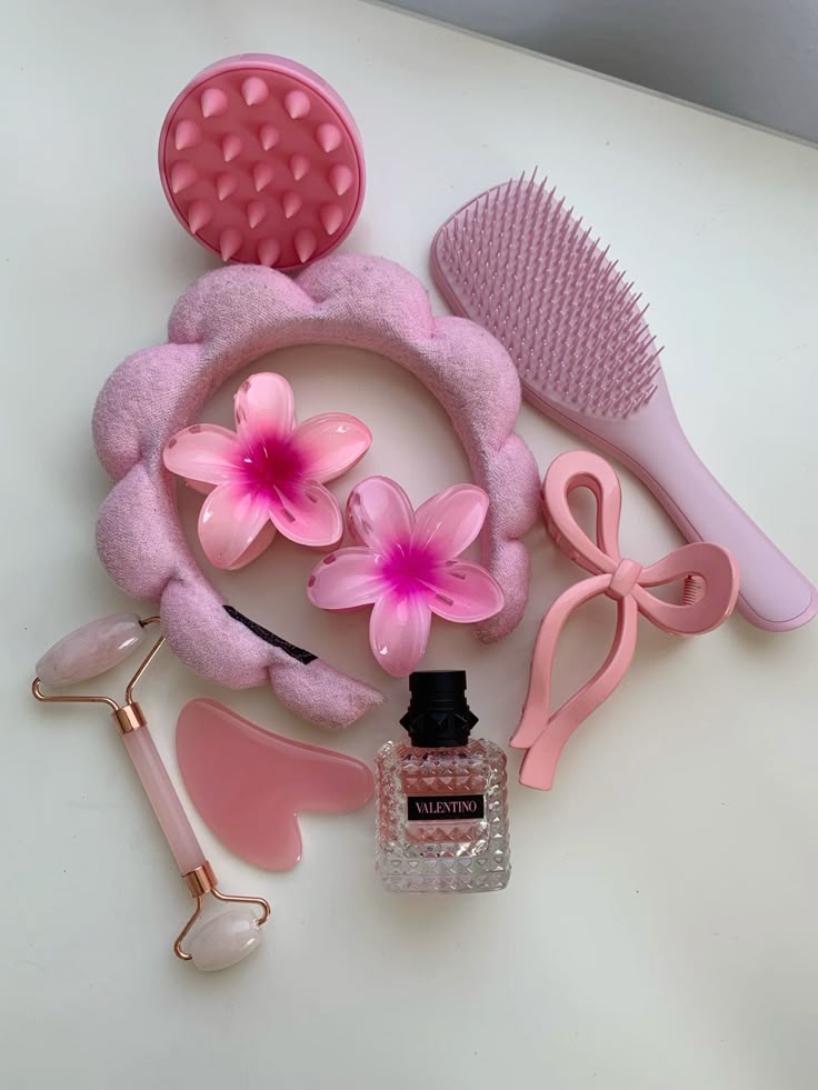 Pink self-care essentials: hairbrush, floral clips, headband, facial roller, gua sha, and perfume bottle on a white background.
