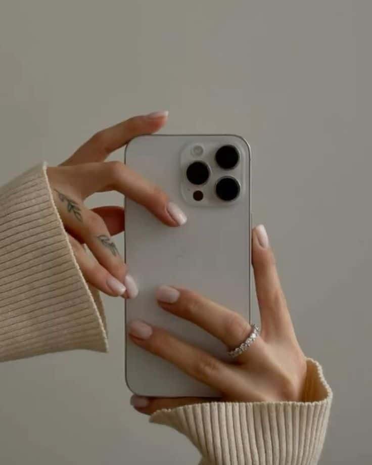 Person with elegant ring and tattoos holding a modern smartphone, focusing on its triple-camera setup. Neutral background.