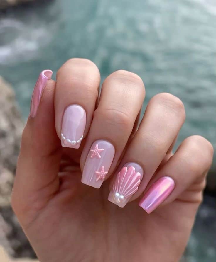 Pink seashell and starfish nail art with pearl accents, set against a scenic ocean view. Perfect for summer beach vibes.