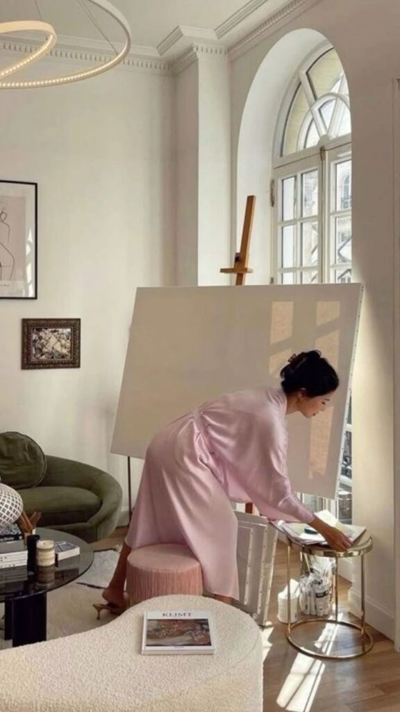Person in a pink robe setting up a creative studio space by a sunlit window, with a blank canvas and cozy decor.