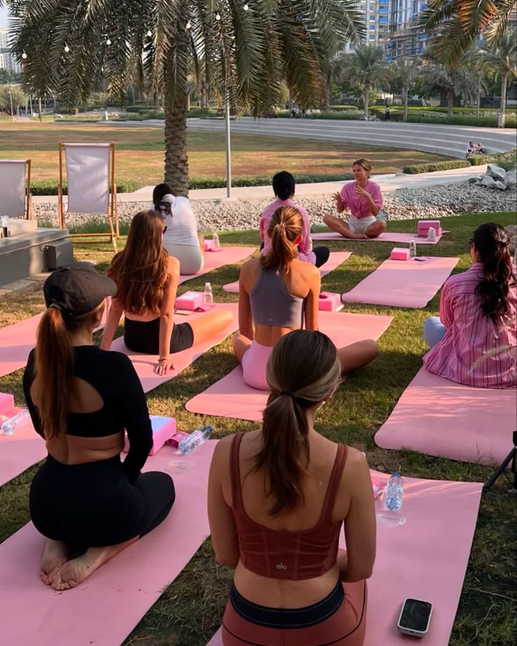 Outdoor yoga class with instructor under palm trees; group practicing meditation on pink mats in a park setting.