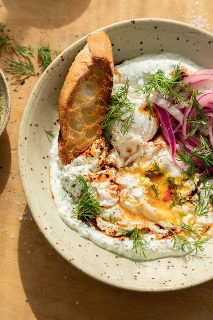 Creamy poached eggs on yogurt with dill, red onions, and crusty bread in a ceramic bowl. Perfect brunch dish with fresh herbs.