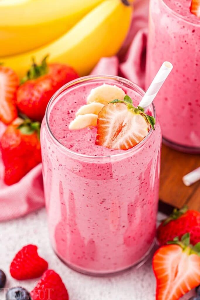 Strawberry Banana Smoothie Recipe - Mom On Timeout - Handy Home Creamy strawberry banana smoothie in a glass, topped with sliced fruit, surrounded by fresh strawberries and bananas.