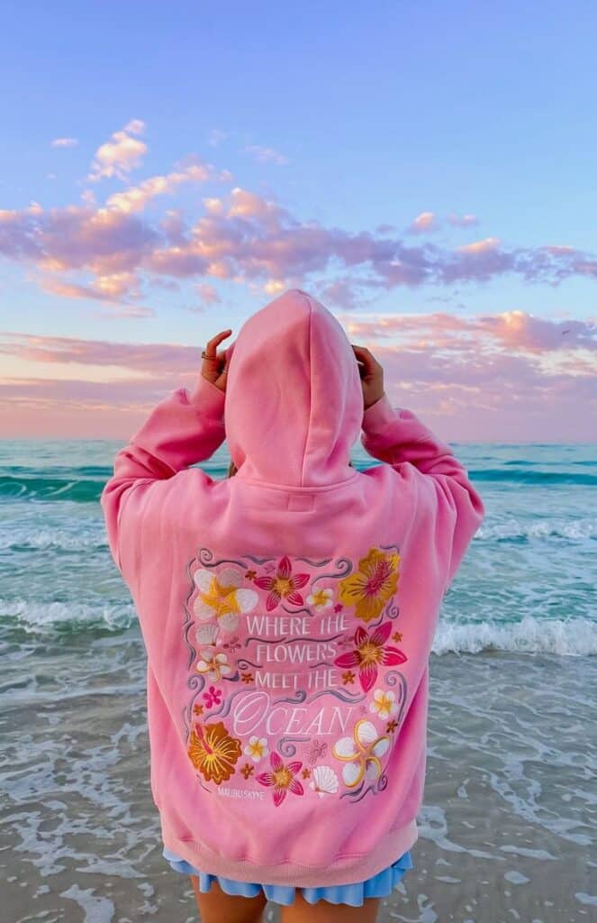 Person in pink hoodie with floral design enjoys ocean view at sunset. Pink sky and gentle waves create a serene beach scene.