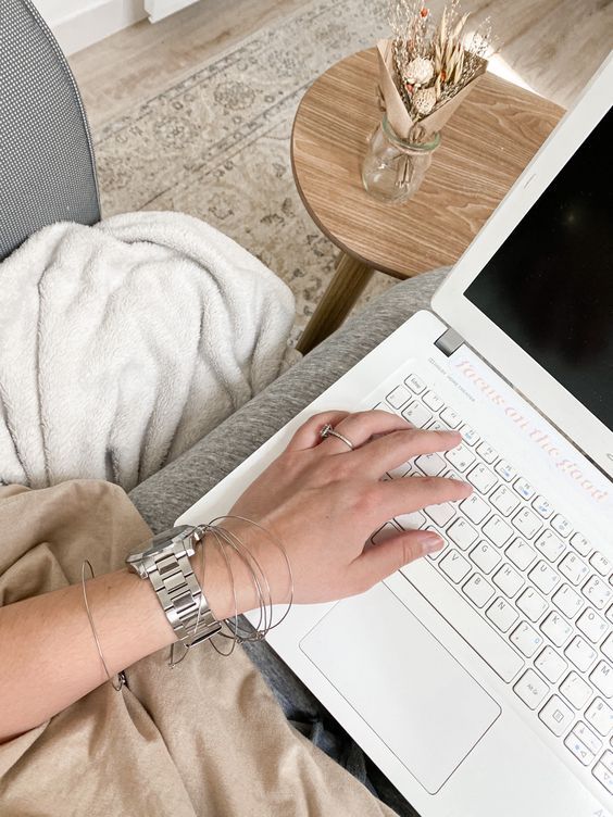 Person typing on a laptop in a cozy home setting, wearing a watch and bracelets. Beige blanket and vase with dried flowers nearby.