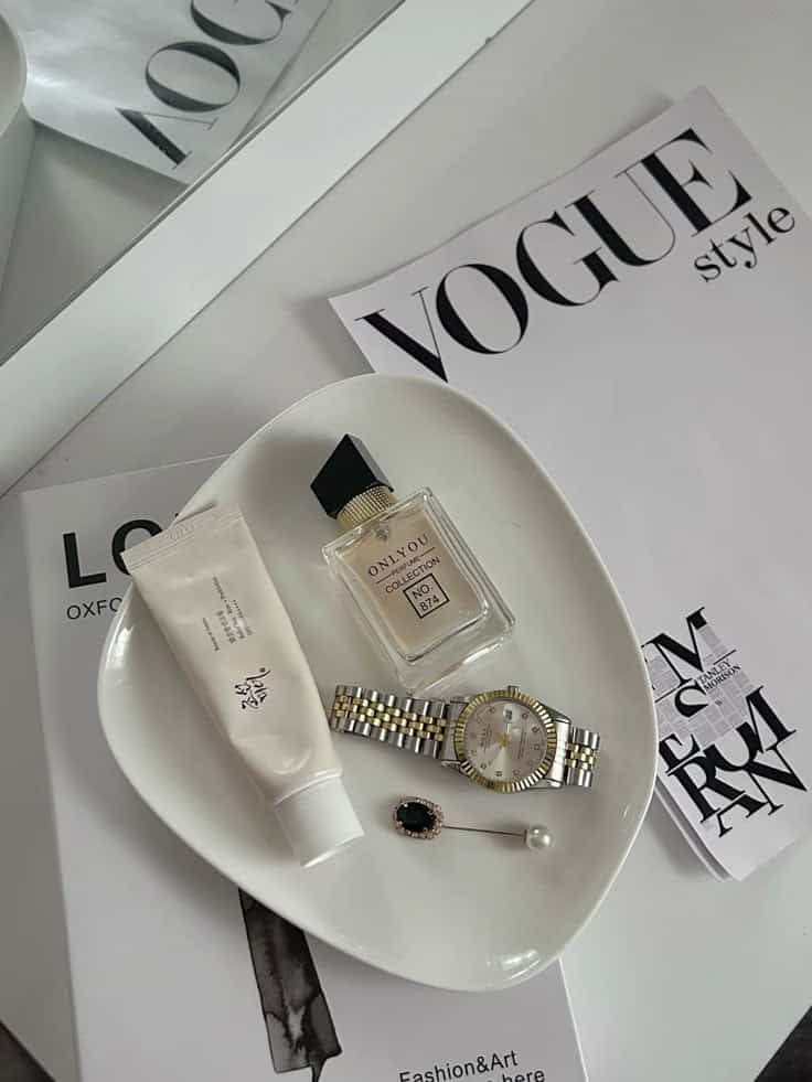 Flat lay of beauty and fashion items on Vogue magazine, includes perfume, cream tube, watch, and brooch on a white dish.