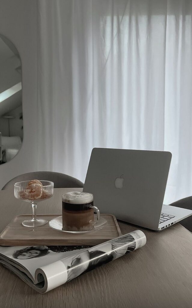 Cozy home office setup with a laptop, coffee, pastry, and magazine on a wooden table by a window with sheer curtains.