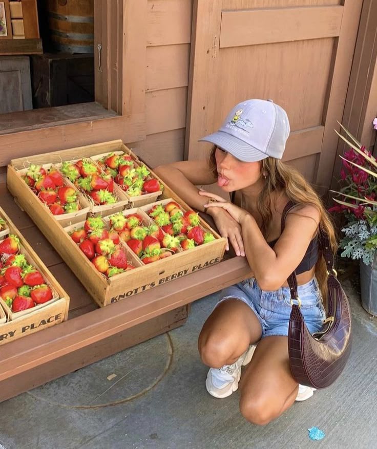 Woman posing playfully by a wooden stand of fresh strawberries at Knotts Berry Place, wearing a cap and casual summer outfit.