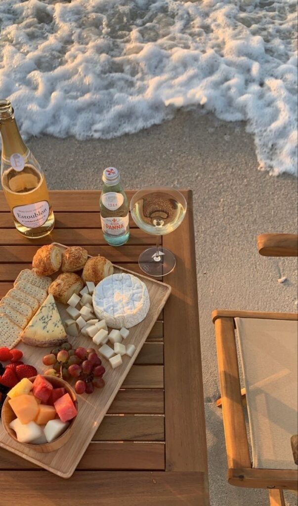 Beachside picnic with cheese, fruit platter, wine, and waves gently crashing near a wooden table setup.