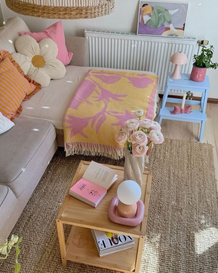 Cozy living room with beige sofa, colorful cushions, floral decor, and a wooden coffee table with books and a lamp.