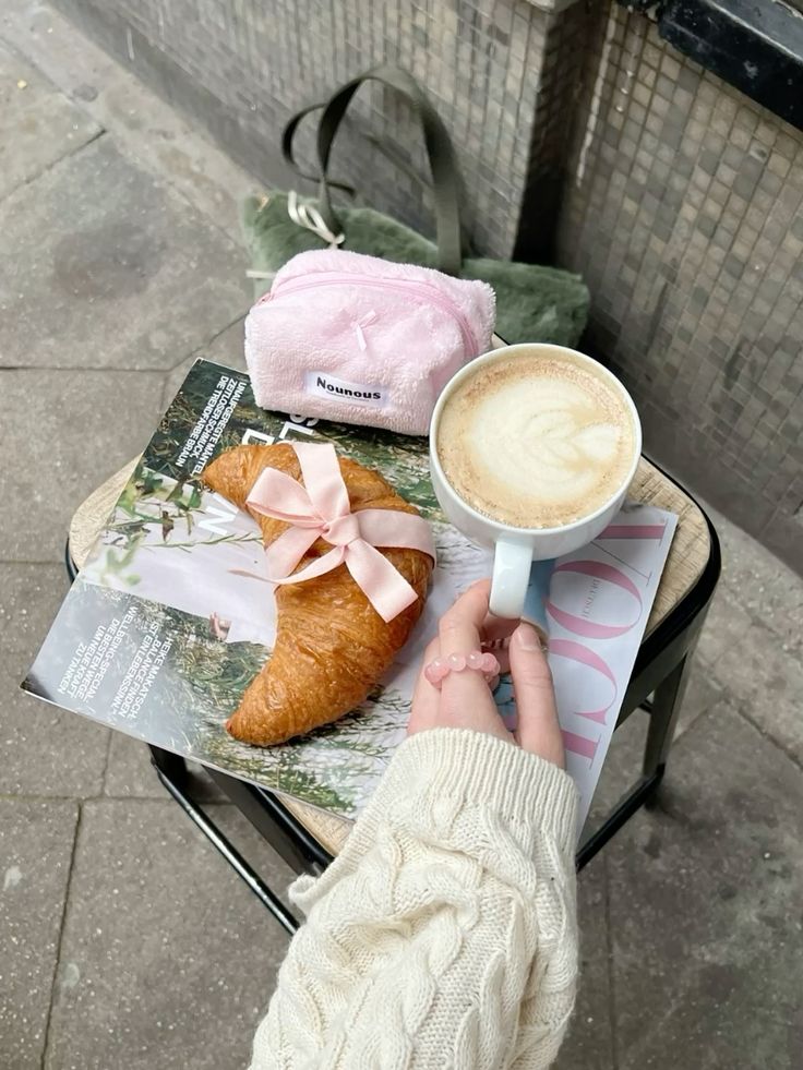 Cozy café vibe: latte, croissant with pink ribbon, and magazine on a small table, perfect for a relaxing morning.