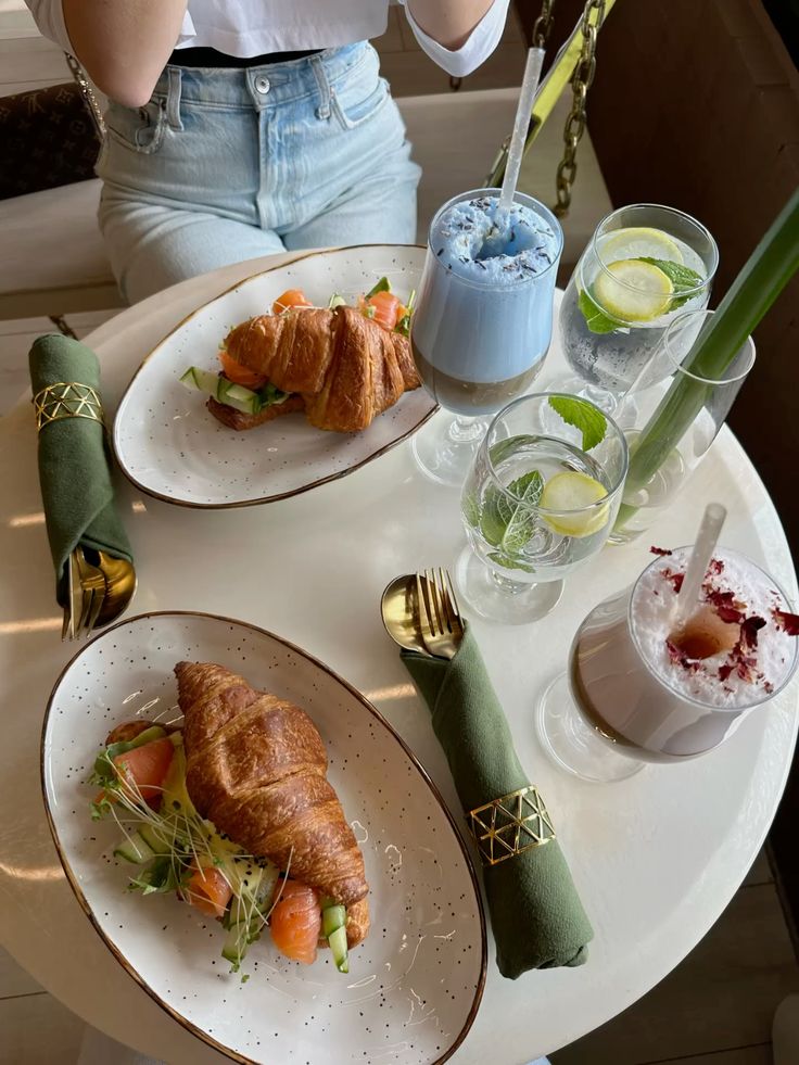 Croissant sandwiches with salmon and avocado, served with vibrant blue smoothie and refreshing lemon mint water on a stylish table.