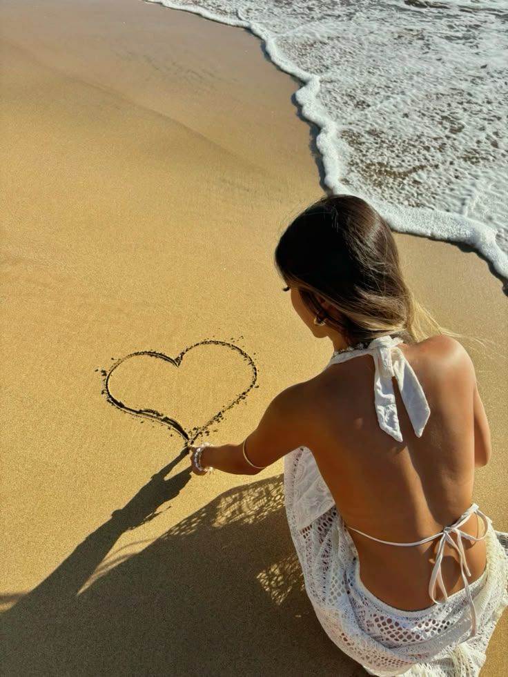 Woman in white dress drawing heart in sand by ocean wave. Romantic beach moment.